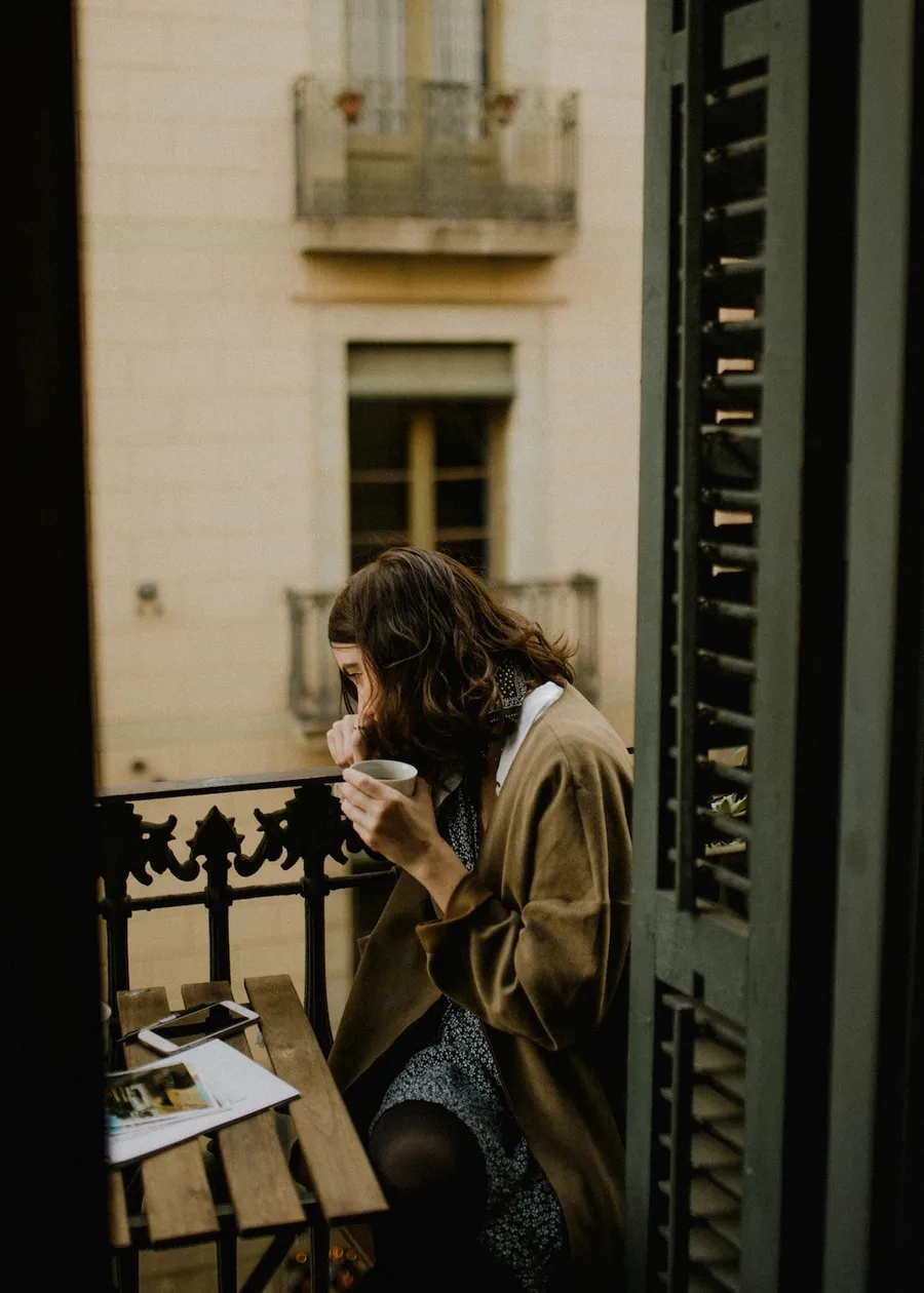 Original photo: a woman sipping coffee on a narrow balcony between dark wooden shutters